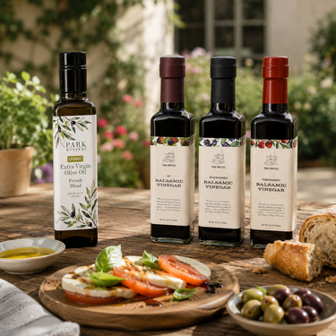 Bottles of Park Street Pantry olive oil and vinegar on a wooden table with a plate of food outdoors.