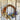 A rustic dried flower wreath hanging on a weathered white wooden door. The wreath features a natural brown grapevine base decorated with orange marigolds, blue lavender, beige grasses, and red rose buds.