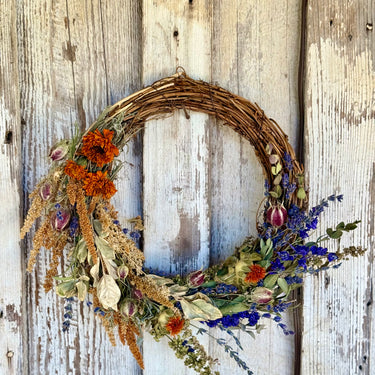 A rustic dried flower wreath hanging on a weathered white wooden door. The wreath features a natural brown grapevine base decorated with orange marigolds, blue lavender, beige grasses, and red rose buds.